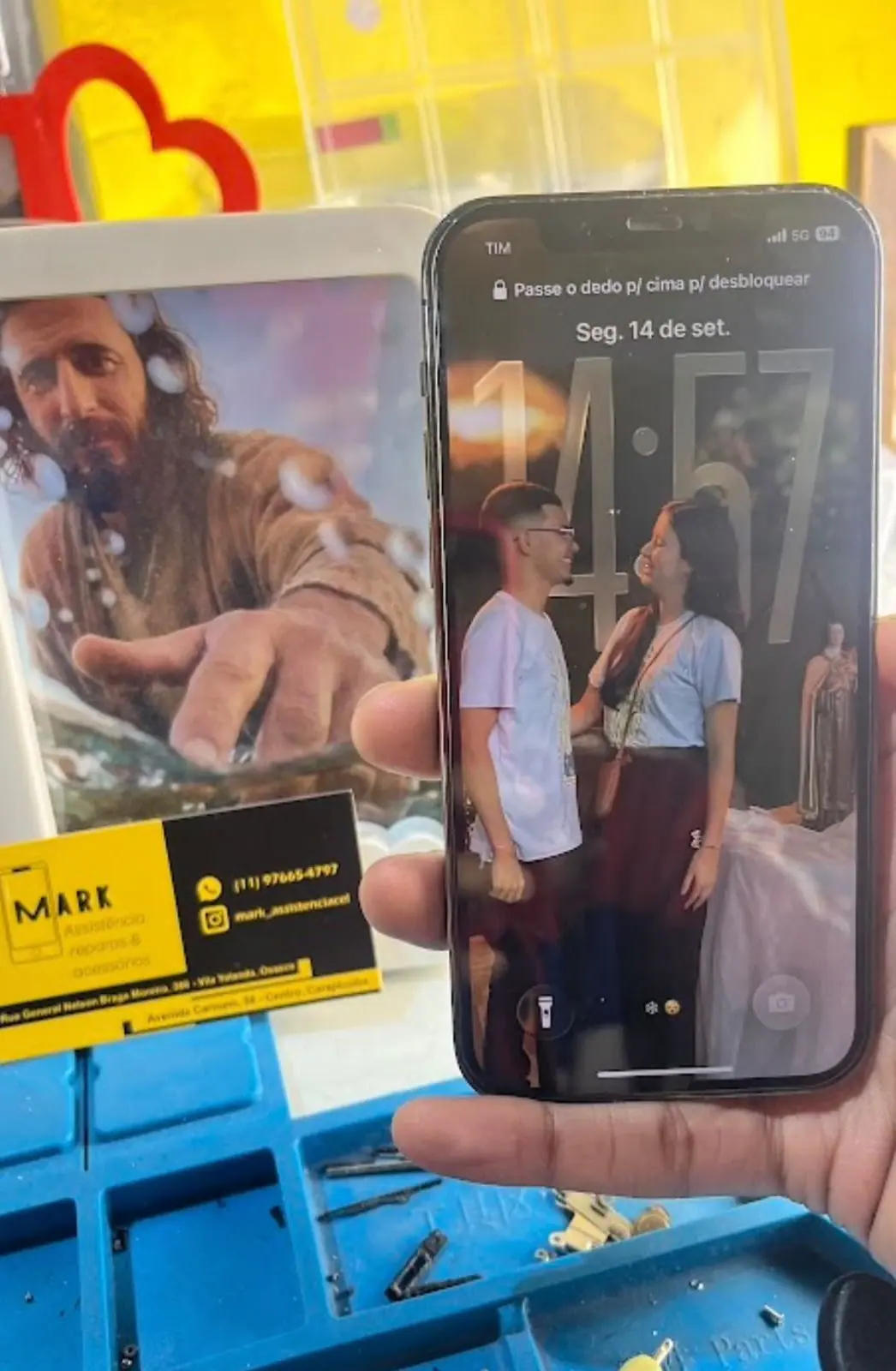iPhone com a tela ligada, segurado diante da bancada de reparo, com o cartão de visita da MARK ao lado.