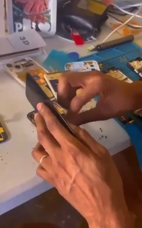 Mãos trabalhando em um iPhone na bancada, entre peças e ferramentas.
