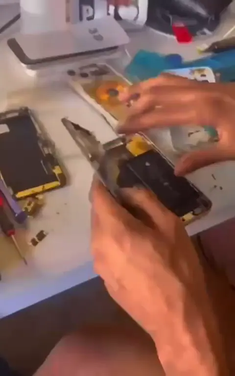 Mãos trabalhando em um iPhone aberto na bancada, com peças e ferramentas ao redor.