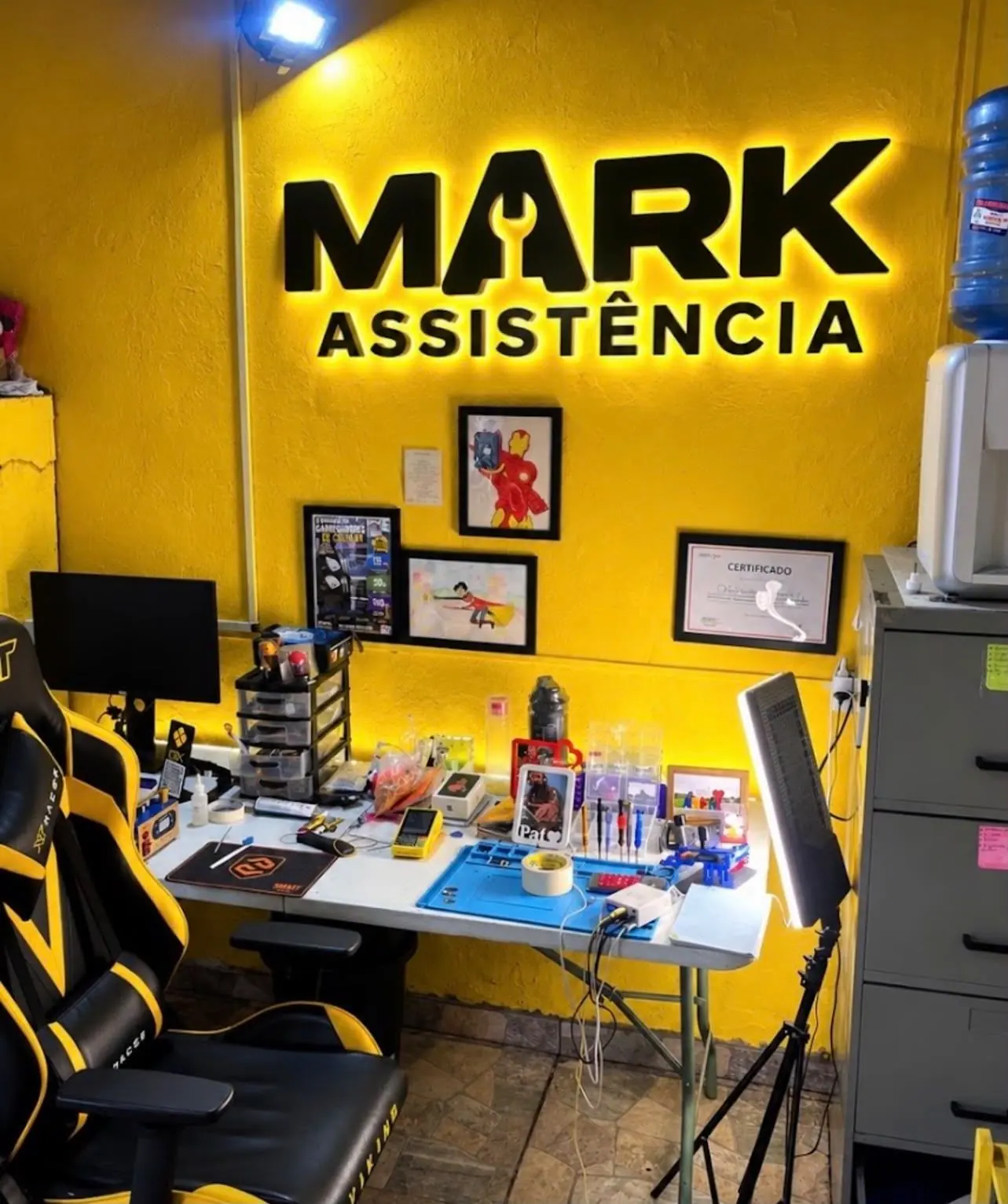 Interior da loja da MARK ASSISTÊNCIA: parede amarela com o letreiro iluminado da marca, bancada de reparo com ferramentas e cadeira de trabalho.